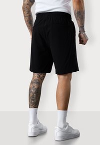 Black shorts with an elastic waistband, featuring a back pocket. Worn with white Nike sneakers and white crew socks. Exposed tattoos are visible.