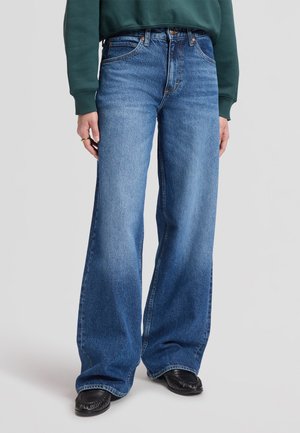 Person iført blå jeans med høj talje og brede ben, kombineret med sorte sko og en grøn sweatshirt, står mod en ensfarvet baggrund.