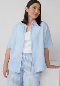 Lichtblauwe kortslurf knoopshirt in een linnenmix. Heeft een zachte textuur en een casual pasvorm. Gemaakt over een witte tanktop en gestreepte broeken.