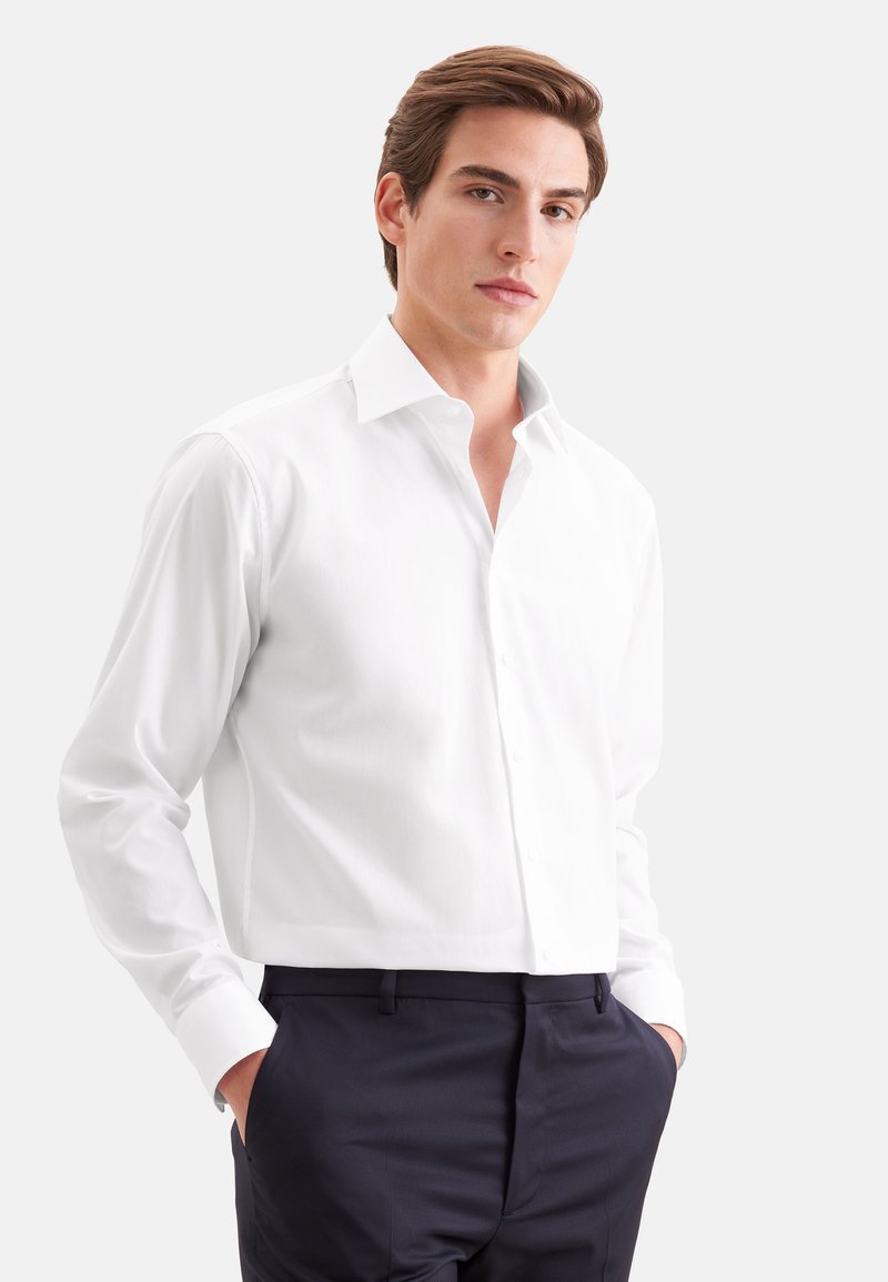 Homme debout, les mains dans les poches, portant une chemise blanche et un pantalon foncé sur un fond clair uni.