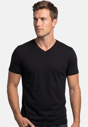 Junger Mann mit kurzen braunen Haaren, der ein eng anliegendes schwarzes V-Ausschnitt-T-Shirt trägt und leicht nach links schaut vor einem schlichten hellen Hintergrund.