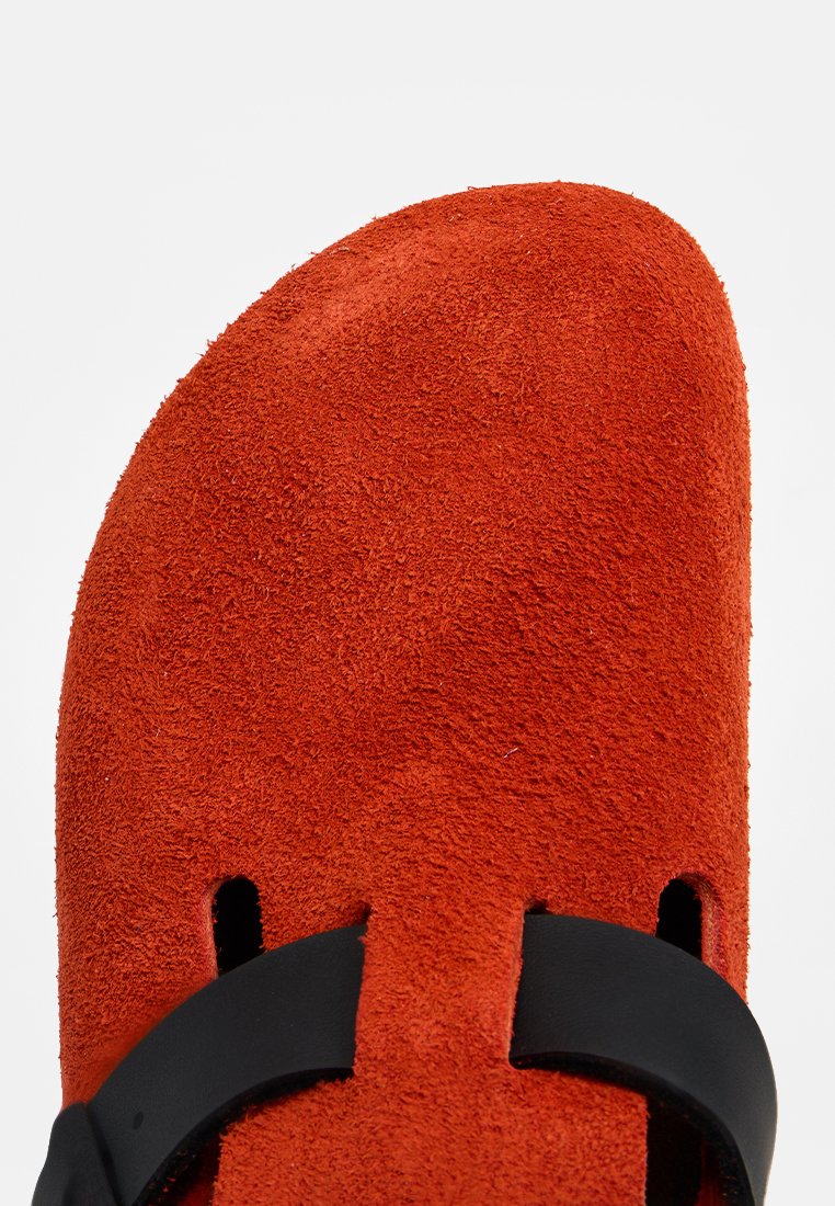 Nahaufnahme der roten Wildleder-Schuhspitze mit schwarzem Lederriemendetail vor einem weißen Hintergrund.