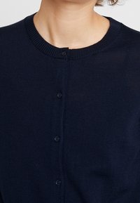 Cardigan lavorato a maglia blu navy con scollo rotondo, dotato di cinque bottoni davanti. Texture morbida con bordi a coste attorno al collo e ai polsini.