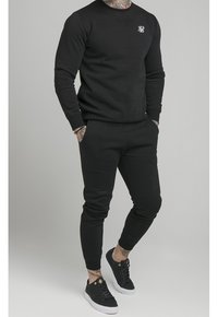 Černá bavlněná souprava mikiny a tepláků s kulatým výstřihem, žebrovanými manžetami a malým logem. Kombinováno s černými teniskami.