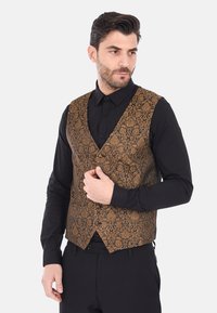 Gilet marrone a fantasia con rever a punta, realizzato in tessuto testurizzato, indossato sopra una camicia nera a maniche lunghe, con un design aderente e tasche laterali.