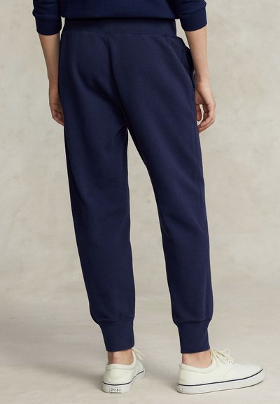 Άτομο που φοράει ναυτικό μπλε παντελόνι τζόγκινγκ και λευκά αθλητικά παπούτσια, στέκεται με τα χέρια χαλαρά στο πλάι μπροστά σε μια απλή φόντο.