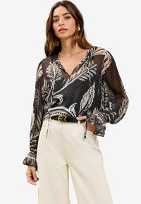 Bluză neagră din material transparent cu modele paisley și puncte albe; are un gât în V, șnururi de reglare și mâneci lungi cu volane.