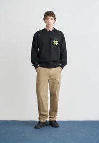 Felpa nera con grafica di trifoglio verde e scritte, abbinata a pantaloni cargo beige con tasche laterali e vestibilità comoda. Scarpe da ginnastica nere.