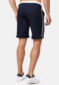 Marineblå shorts med en jevn tekstur, oppbrettede ermer og hvite side stripe. Har baklommer med knappelukking.