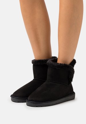 Jambes portant des bottes d'hiver noires montant jusqu'à la cheville avec une doublure en fourrure moelleuse, debout sur une surface blanche.