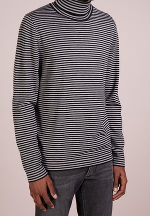 Personne portant un pull à col roulé rayé horizontal noir et gris avec un jean gris foncé sur un fond uni.