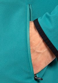 Türkiser Blazer mit schwarzen Akzenten, ausgestattet mit einer Reißverschlusstasche. Nahaufnahme zeigt die Textur des Stoffes und eine Hand, die in der Tasche ruht.