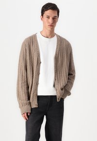 Beige kötött kardigán texturált mintával, nyitott elejű, öt fa gombbal. Fehér, kerek nyakú pólóval és sötét farmernadrággal párosítva.