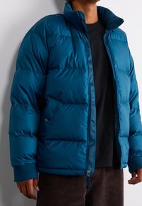 Grönblå puffjacka med hög krage, dragkedja framtill och ribbade ärmslut. Har sydda paneler och en glänsande yta. Bärs över en svart skjorta.