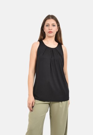 Donna con capelli lunghi che indossa un top nero senza maniche e pantaloni beige, in piedi davanti a uno sfondo chiaro e uniforme.