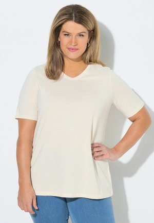 Mujer con cabello castaño claro hasta los hombros, vistiendo una camiseta crema de manga corta con cuello en V y vaqueros azules, posando con la mano en la cadera.