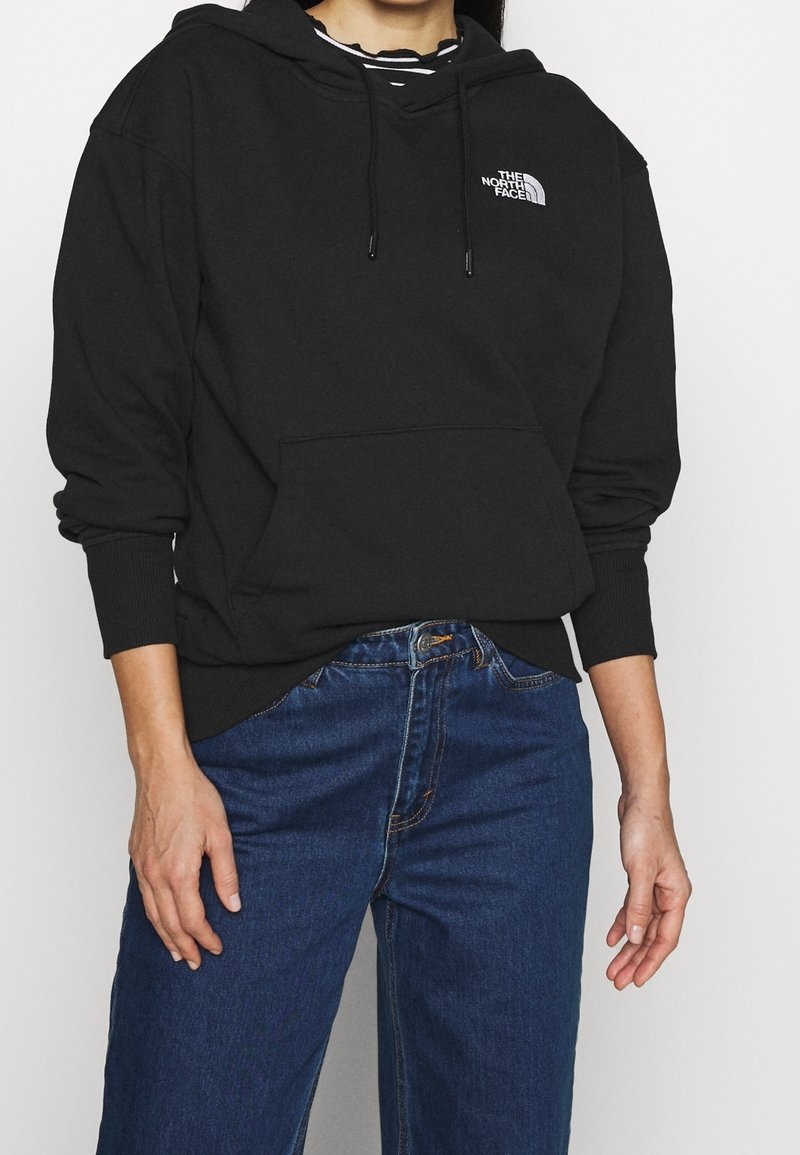 Personne portant un sweat à capuche noir The North Face par-dessus une chemise rayée associée à un jean bleu taille haute, debout les mains détendues.