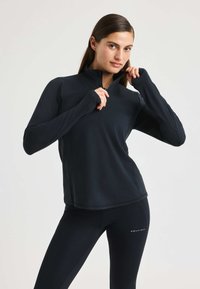Camiseta atlética negra de manga larga con cuello de media cremallera, hecha de una tela elástica, con orificios para los pulgares en los puños y una textura suave.