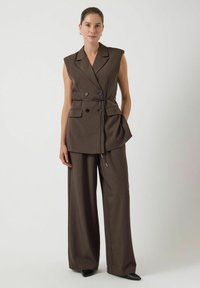 Bruine mouwloze vest met een dubbelbreasted ontwerp, een ceintuur in de taille en twee zakken aan de voorkant, gecombineerd met broek met wijde pijpen.