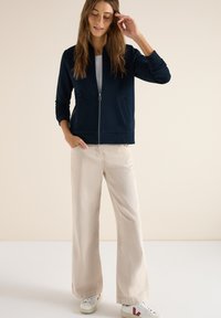 Femme portant une veste zippée bleu marine, un haut blanc, un pantalon large beige et des baskets blanches avec des accents rouges, debout devant un fond beige.