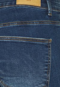 Jeans de mezclilla azul oscuro con costuras contrastantes en color naranja, un bolsillo trasero y una etiqueta de cuero marrón claro en la cintura.