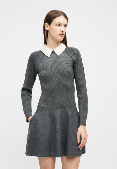 Femme portant une robe grise côtelée à manches longues avec une jupe évasée et un col pointu blanc texturé, debout les mains dans les poches.