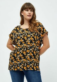 Sort blomsterbluse med gule og hvide blomster, korte ærmer og en afslappet pasform. Sammen med blå jeans. Glat stofstruktur.
