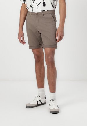 Braune Baumwollshorts mit umgeschlagenen Bündchen, ausgestattet mit einer flachen Vorderseite und einem Knopfverschluss. Kombiniert mit einem gemusterten Hemd und lässigen Sneakers.