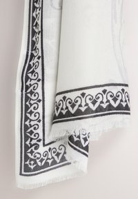 Tissu blanc avec motif de bordure noire ornée, plié avec des bords frangés, suspendu sur un fond clair.