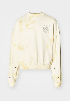 Krémová mikina so žltými tie-dye vzormi, okrúhly výstrih, rebrované manžety a vyšívané písmeno 'A' na hrudi.