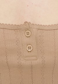 Κοντινό πλάνο μπεζ πλεκτού τοπ με κάθετα σχέδια δαντέλας και δύο μπεζ κουμπιά κοντά στη σκαλοειδή λαιμόκοψη πάνω σε γυμνό δέρμα.