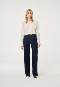 Crème, textuur, blouse met lange mouwen en knoopdetail, voorzien van borduursel, gecombineerd met donkerblauwe, losse spijkerbroek. Het schoeisel heeft een bedrukt ontwerp.