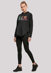 Sudadera negra con un logo colorido de Marvel, corte relajado, con aberturas laterales, combinada con leggings negros y zapatillas negras. Textura de algodón suave.