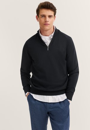 Mann in einem schwarzen Halbzip-Pullover über einem weißen Hemd und blauen Hosen, der mit Händen in den Taschen vor einem neutralen Hintergrund steht.