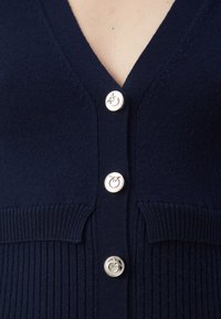 Navy gebreide cardigan met V-hals, geribbelde textuur aan de zoom en zilveren knopen met een ronde vorm. Belangrijk detail: verticale patroon.