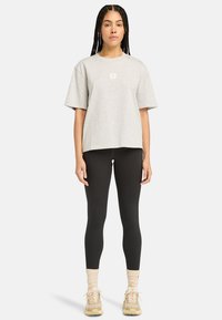 Šedé oversize tričko s malým logem na hrudi, spárované s černými legínami a béžovými sportovními botami, na nohou s ribbovanými krémovými ponožkami.