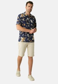 Korte mouwen blauwe shirt met wit en oranje tropisch bladmuster, gecombineerd met beige opgerolde shorts en beige instappers.