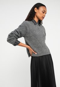 Giovane donna che indossa un maglione grigio a uncinetto sopra una camicia a righe bianco e nero, abbinato a una lunga gonna plissettata nera, in posa di profilo.