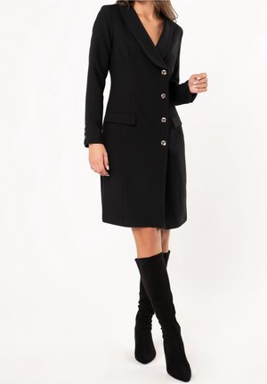 Vrouw draagt een zwarte blazerjurk met knopen en klepzakken, en zwarte suède kniehoge laarzen, staand tegen een witte achtergrond.