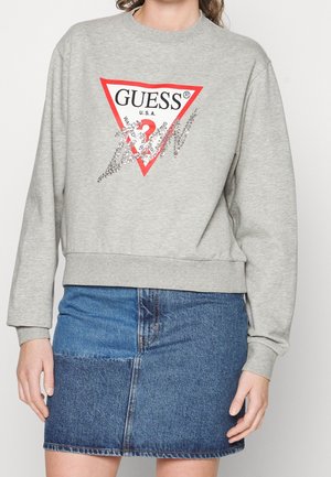 Personne portant un sweat-shirt gris avec un logo Guess rouge et blanc et une mini-jupe en denim patchwork bleu.