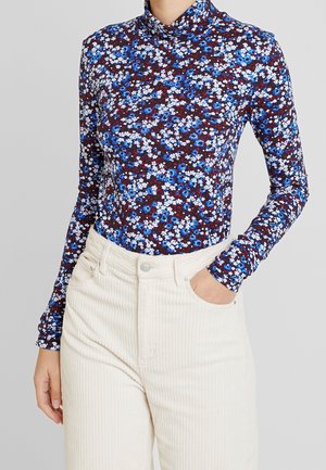 Kobieta ubrana w dopasowany, długi rękaw z golfem w niebieski, biały i bordowy wzór kwiatowy, włożony w beżowe, wysokiej talii sztruksowe spodnie.