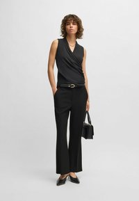 Zwarte mouwloze top met wikkeldesign, gecombineerd met hoge zwarte broek. Gladmateriaal, aansluitende silhoehette en wijde pijpen. Houdt een kleine zwarte tas vast.