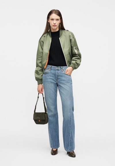 Γυναίκα που φοράει ένα χακί μπουφάν τύπου μπομβέρ πάνω από μαύρο τοπ, ανοιχτό μπλε τζιν με ψηλή μέση, καφέ ψηλοτάκουνα και κρατά μια μικρή σκούρα πράσινη τσάντα χειρός.