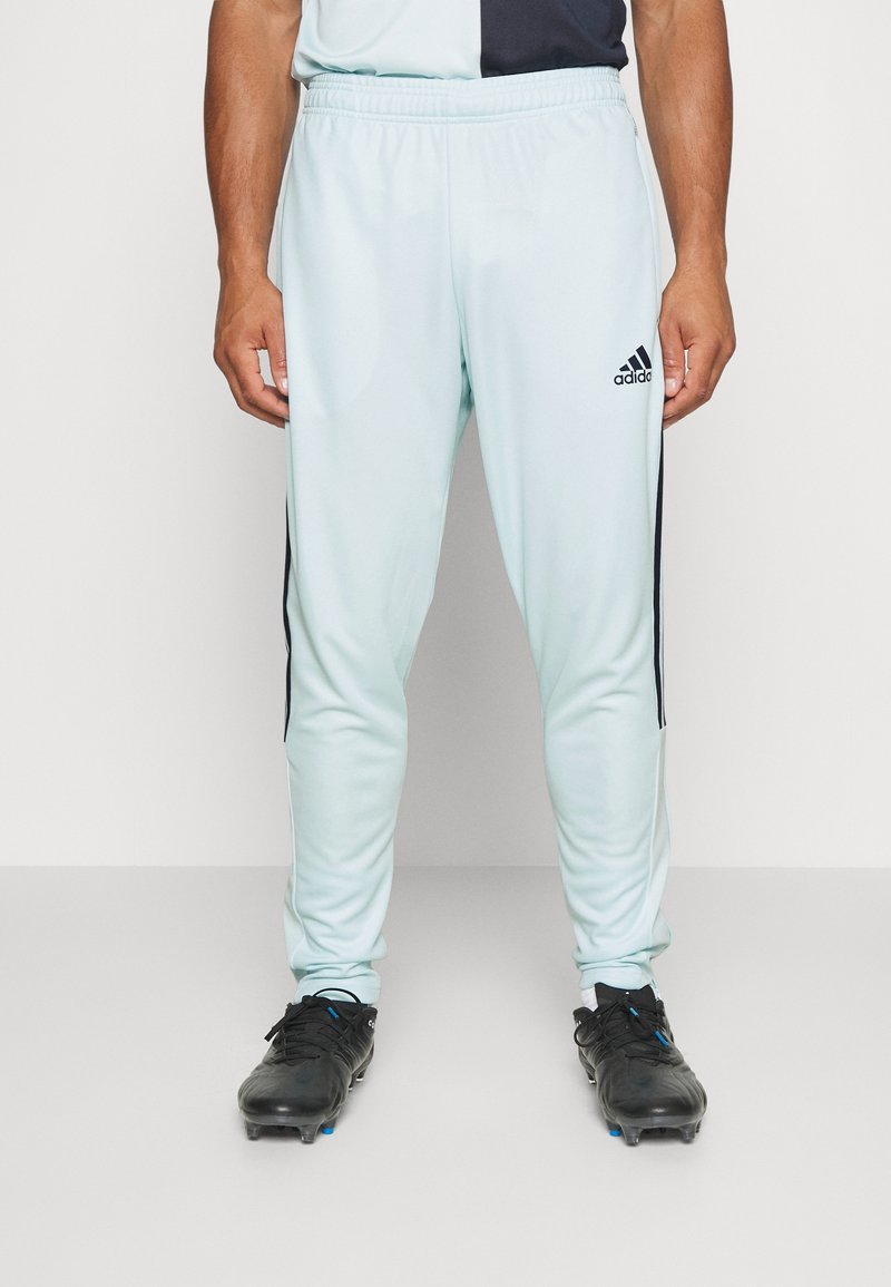 Helles blaues Sporthose mit schwarzen Seitenstreifen und elastischem Bund, kombiniert mit schwarzen Fußballschuhen. Das Material scheint glatt und dehnbar zu sein.