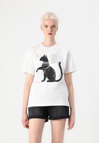 Hvit bomull t-skjorte med et grafisk motiv av en svart katt prydet med perlelignende halskjeder. Designet inkluderer tekst ved siden av illustrasjonen.