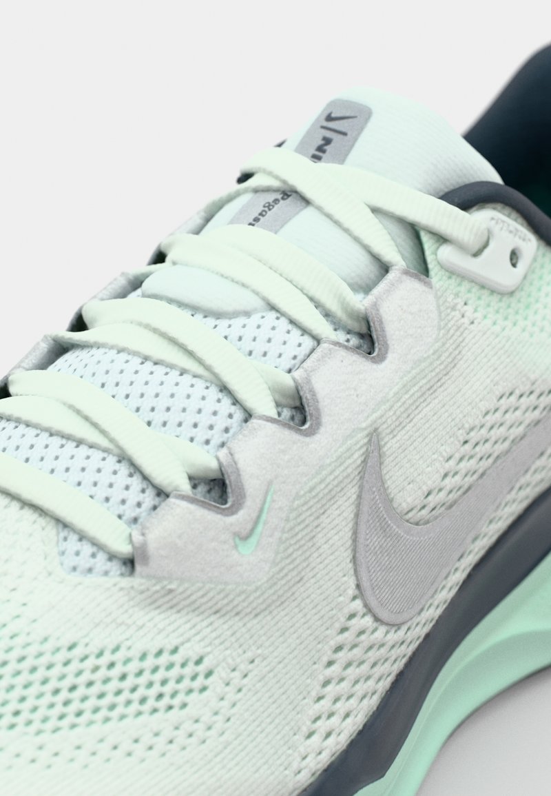 Zapatilla deportiva en verde claro y gris con textura de malla, cordones planos y un logo de Swoosh reflectante. Cuenta con una suela acolchada y puntera reforzada.