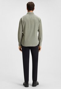 Man die met zijn rug naar de camera staat, gekleed in een lichtgroen overhemd met lange mouwen, zwarte broek en zwarte schoenen, tegen een witte achtergrond.