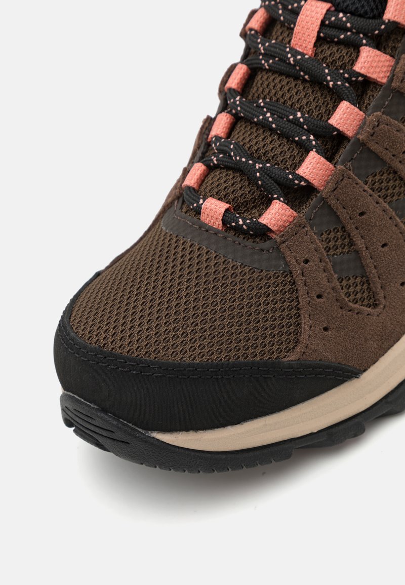 Zbliżenie brązowego sportowego buta z siateczkową cholewką, czarnymi i różowymi sznurówkami, zamszowymi detalami, beżową podeszwą środkową i czarną gumową podeszwą.