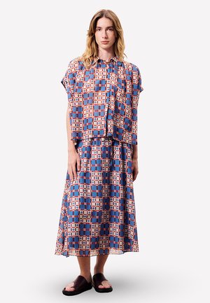 Femme portant un chemisier ample à carreaux bleus et orange avec une jupe longue assortie, associée à des sandales ouvertes brun foncé, debout devant un fond blanc.