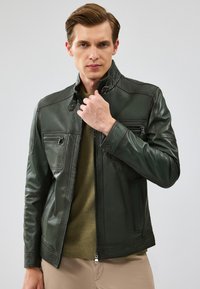 Giacca in pelle verde scuro con zip frontale, collo alto, tasche sul petto con zip e tasche laterali. Presenta dettagli cuciti e una texture liscia.